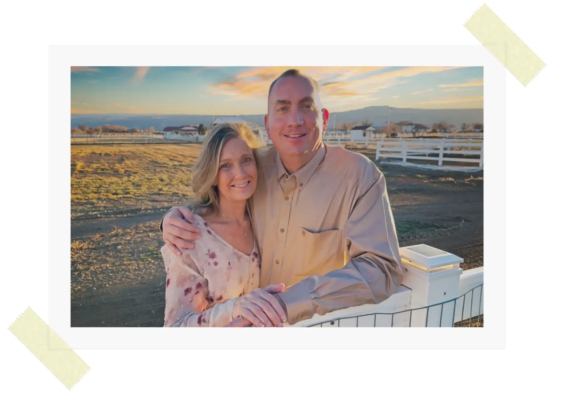 A couple smiles and poses together outdoors by a white fence, with a sunset sky and open field in the background. The photo has a white border and is taped at two corners.